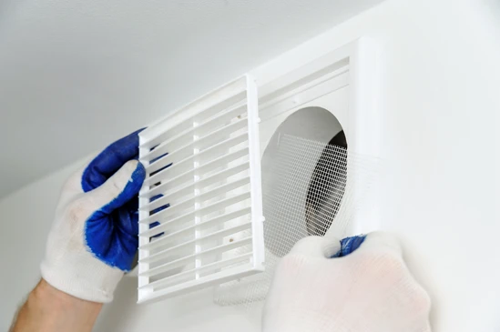 Intervention de maintenance dans un logement, technicien portant des gants de protection bleus et blancs procédant au démontage d'une grille de ventilation murale blanche