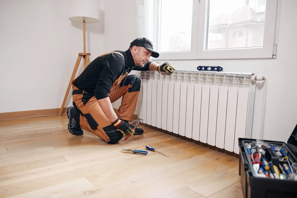 Plombier professionnel en combinaison de travail installant ou réparant un radiateur à ailettes blanc dans un appartement moderne