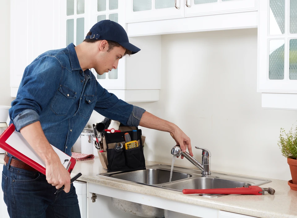 Plombier en combinaison bleue et casquette répare un robinet de cuisine dans une maison moderne