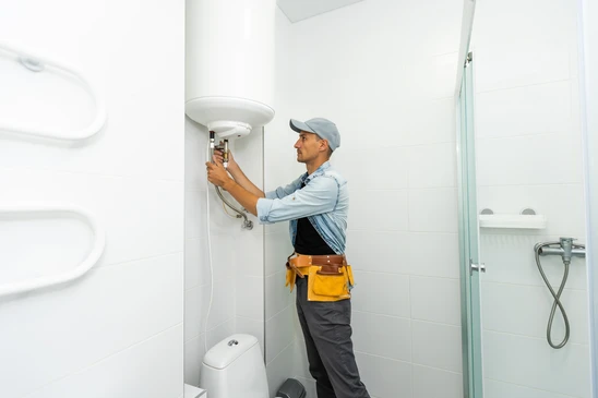 Plombier professionnel en salopette et casquette installant ou réparant un chauffe-eau électrique mural dans une salle de bain moderne